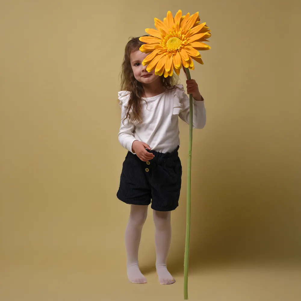 Séance photo d'une petite fille en studio, tenant une grande fleur jaune et posant devant un fond jaune pastel.