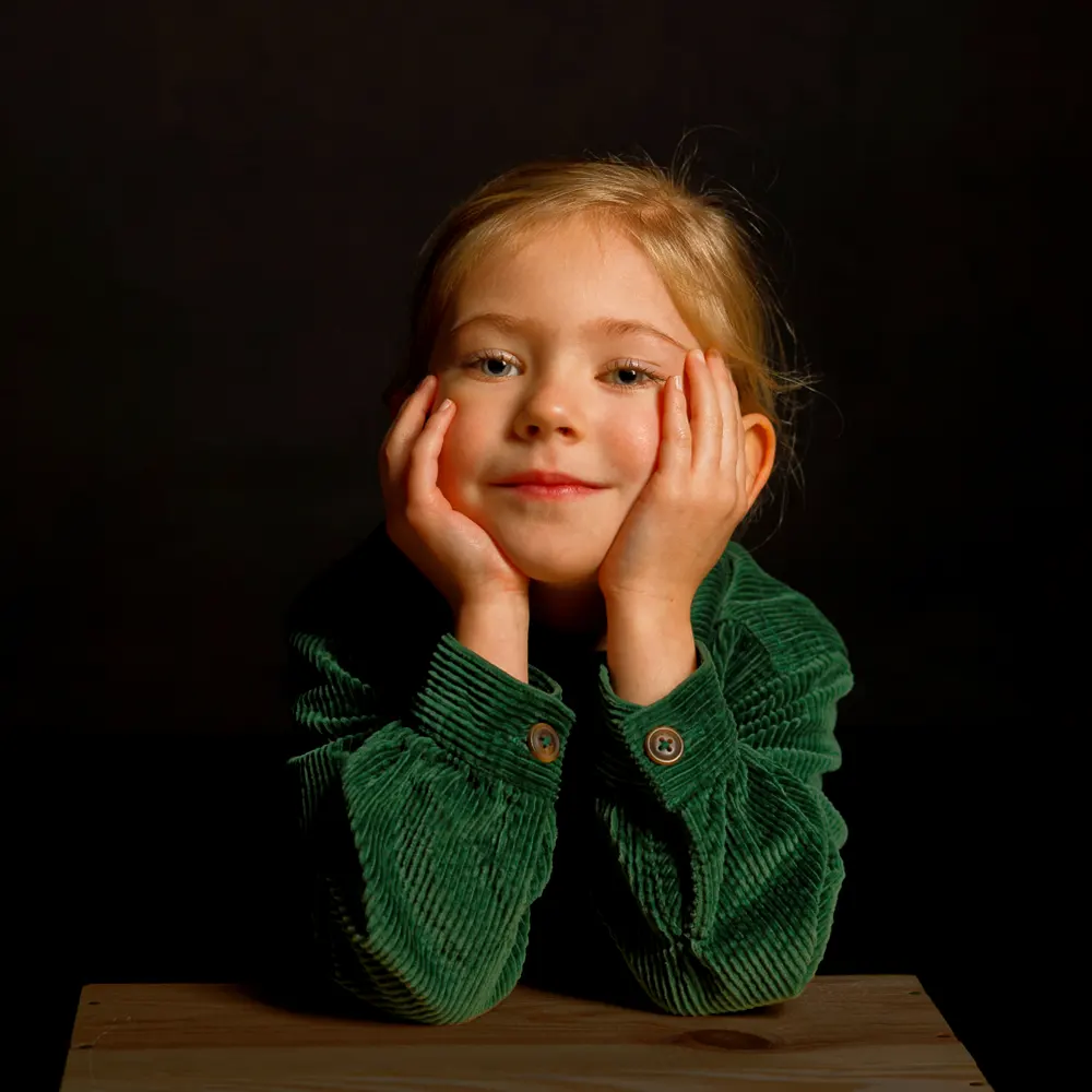 Portrait studio d'une petite fille avec les mains sous le menton, sur un fond sombre.