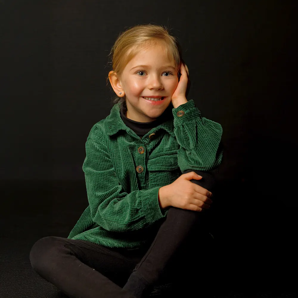 Portrait studio d'une petite fille souriante, assise avec les mains sous le menton, sur un fond sombre.