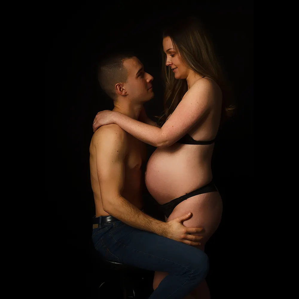 Portrait de grossesse en studio pour un couple sur fond noir. Le futur papa est assis sur un tabouret, la future maman est debout, ils se regardent tendrement en tenant son ventre.