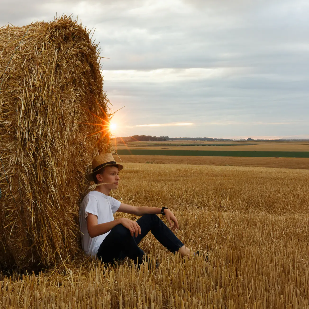 Portrait d'un jeune homme assis contre une botte de foin au coucher du soleil, pour une séance photo en extérieur.