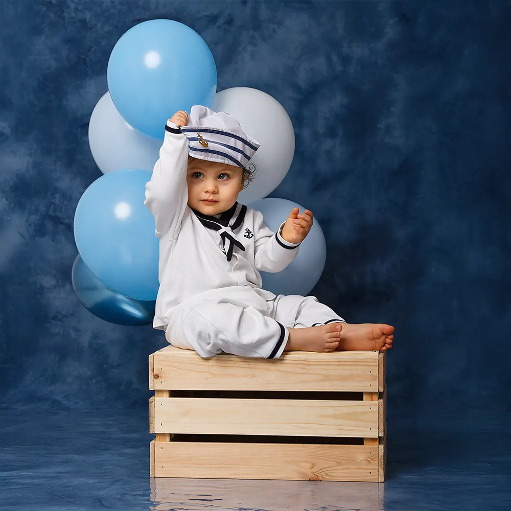 · Séance photo d'anniversaire pour un an, bébé déguisé en matelot, assis sur une caisse en bois avec des ballons bleus.