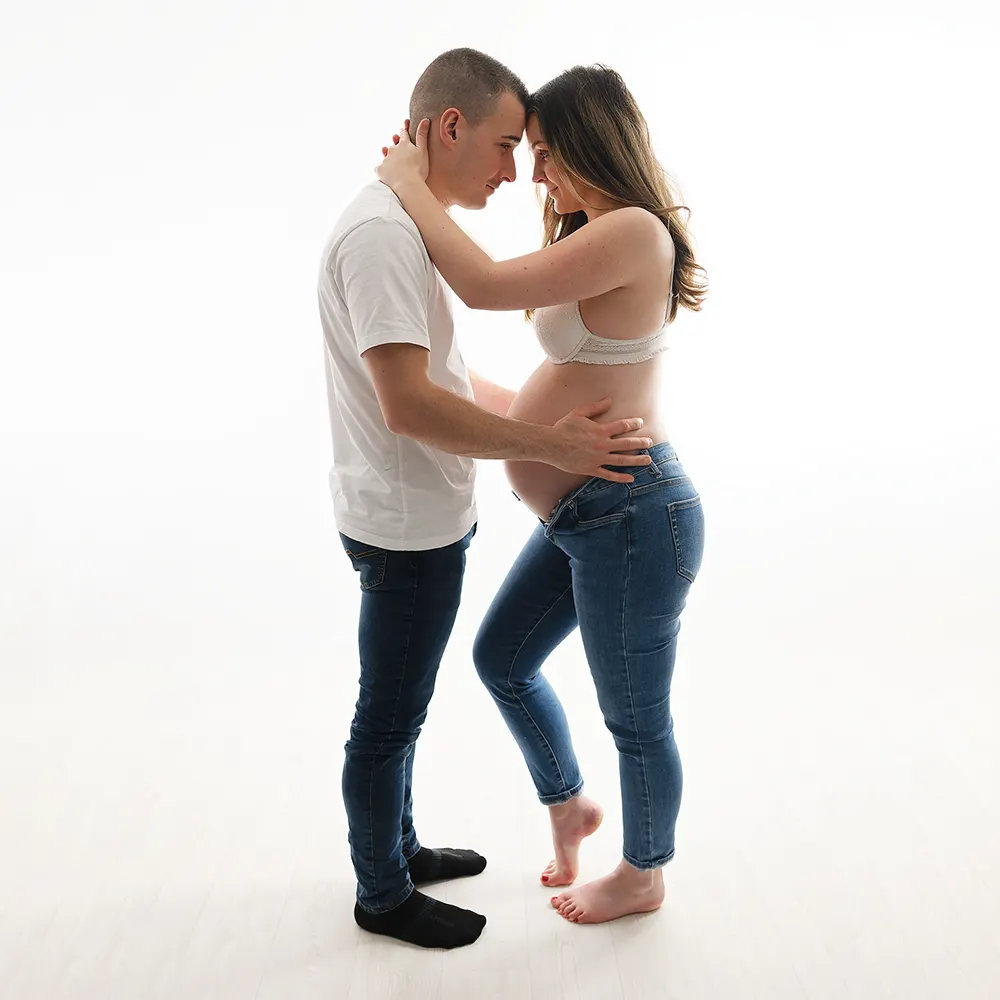  Séance photo de grossesse en studio lumineux. Un couple se tenant tendrement, la future maman en soutien-gorge blanc et jeans ouverts, le futur papa caressant son ventre.