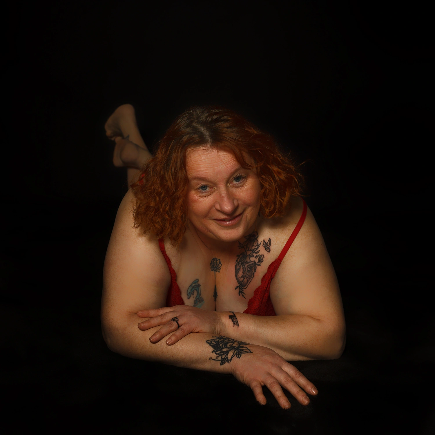 Portrait boudoir d'une femme rousse souriante et confiante, allongée en lingerie rouge. Photo réalisée par votre photographe boudoir à Sainte-Catherine, près d'Arras.