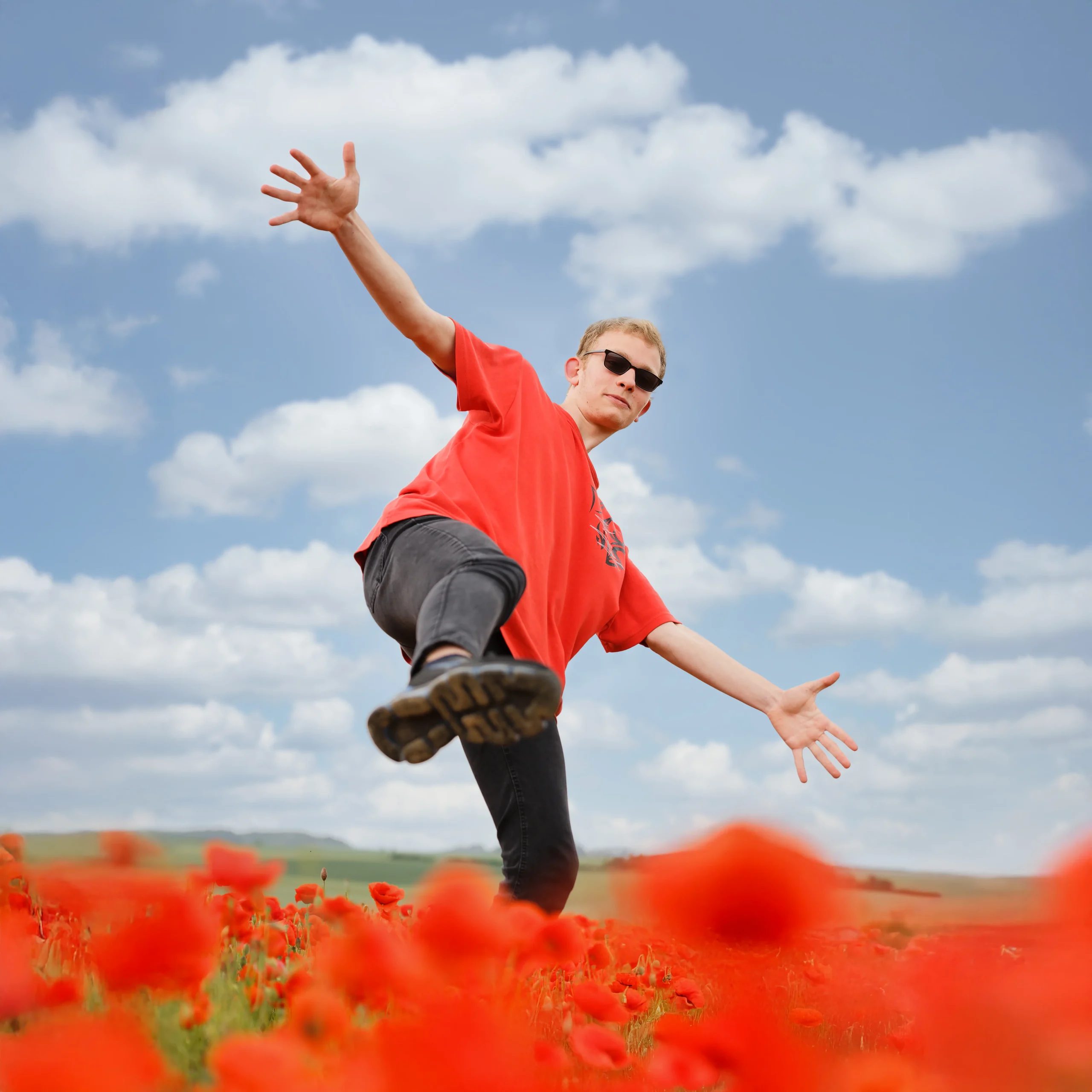 Portrait dynamique de Yannis sautant dans un champ de coquelicots, en t-shirt rouge et lunettes de soleil.